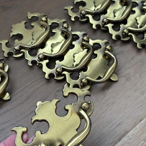 May include: A collection of antique brass drawer pulls. Each pull features an ornate, stylized backplate with a curved handle. The brass has a warm, golden hue, and the design suggests a vintage or classic aesthetic. The pulls are arranged on a wooden surface.