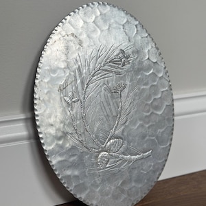 May include: An oval-shaped silver-toned metal tray featuring a hammered texture across its surface. The center of the tray displays an embossed design of a pine branch with needles and two pine cones. The edges of the tray have a scalloped border.