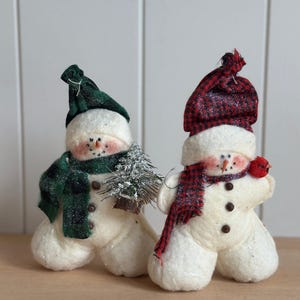 May include: Two plush snowman figurines with white textured bodies, brown button accents, and orange carrot noses. One wears a green plaid hat and scarf while holding a small frosted pine tree. The other wears a red plaid hat and scarf with a red bird.
