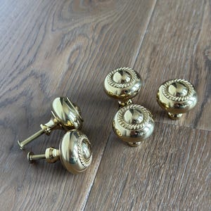 May include: Several shiny, gold-colored door knobs with a decorative rope-like design around the top. The knobs are round and have a screw attached to the back. The knobs are arranged on a light brown wooden surface.