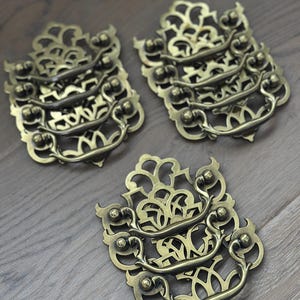 May include: Ornate brass drawer pulls with a decorative filigree design. Each pull features a curved handle and intricate openwork, creating a vintage aesthetic. The pulls are arranged on a wooden surface.