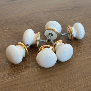 May include: Six white ceramic cabinet knobs with gold-colored trim. The knobs are round with a smooth, glossy finish and are attached to metal screws. The knobs are arranged on a wooden surface.