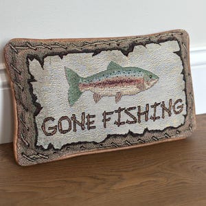 May include: A rectangular woven throw pillow featuring a central image of a trout fish and the text GONE FISHING in dark brown letters. The pillow has a light-colored center panel with a textured border resembling wood grain and a brown trim.