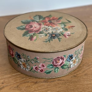 May include: Round, vintage tin box with a floral needlepoint design. The lid features a bouquet of pink, red, and white flowers on a tan background. The sides are adorned with a floral border. The tin has a gold-colored rim.