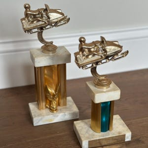May include: Two vintage snowmobile trophies. Each trophy features a gold-colored snowmobile and rider atop a marble base with gold and teal accents. The trophies are displayed on a wooden surface.