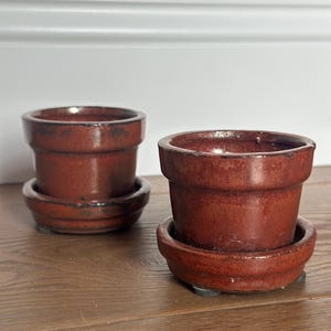 May include: Two small, cylindrical ceramic plant pots with matching saucers. Each pot features a deep, glossy reddish-brown glaze with darker, mottled accents along the rim and base. The pots have a simple, straight-sided design with a wide opening.