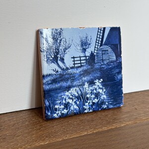 May include: A square ceramic tile featuring a blue and white monochromatic landscape illustration. The scene depicts a windmill, a small wooden fence, trees, and a body of water with foreground vegetation. The tile has a glossy finish and edges.
