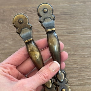 May include: Two antique brass cabinet pulls with a decorative design. The pulls have a warm, aged finish and a classic shape, perfect for adding vintage charm to furniture. Each pull measures approximately 6 inches in length.