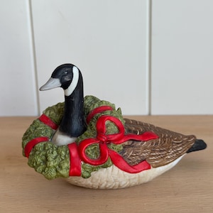 May include: A ceramic Canada goose figurine featuring a black head and neck with a white cheek patch. The goose is wearing a green wreath around its neck, accented with a bright red ribbon tied in a bow. The body has brown and white feathers.
