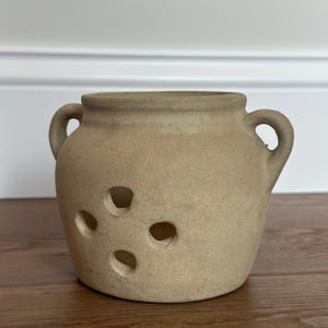May include: A beige, matte-finish ceramic garlic keeper with a rounded body, two side handles, and a wide opening at the top. The front of the vessel features four circular ventilation holes arranged in a diamond pattern to allow for airflow.