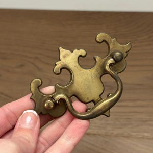 May include: An antique brass drawer pull with an ornate design. The pull features a curved handle and decorative flourishes. The brass has a warm, aged patina, suggesting vintage or antique origins. Ideal for restoring furniture or adding a touch of classic style.