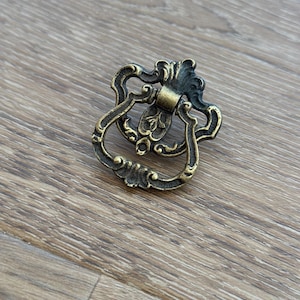 May include: Ornate antique brass drawer pull with a decorative shield-like backplate and a curved handle. The hardware features intricate scrollwork and a warm, aged finish, perfect for vintage furniture restoration projects.