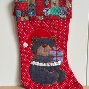 May include: A quilted red Christmas stocking with white polka dots and a cuff featuring a patchwork pattern of holiday icons. An appliqued dark gray bear wearing a red hat holds a blue gift box, accented with a white lace trim at the waist.