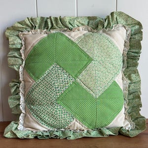May include: A square throw pillow featuring a quilted patchwork design in shades of green and cream. The center displays geometric patterns with floral and polka dot prints, surrounded by a ruffled green floral border and white eyelet trim.