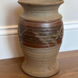 May include: A tall, cylindrical stoneware vase with a flared rim and base. The exterior features a matte, light tan finish on the top and bottom sections, while the center band is glazed in a deep, glossy brown with etched cross-hatch patterns.