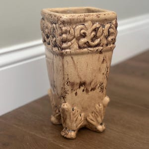 May include: A square ceramic vase with a tan and brown distressed finish. The upper rim features a raised scroll pattern, while the base is supported by four decorative feet with a similar textured design, showing dark brown glaze accents.
