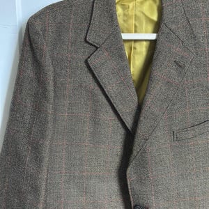 May include: Brown tweed blazer with a red plaid pattern. The blazer has a notched lapel, a single-button closure, and a yellow satin lining. The jacket is hanging on a white hanger.