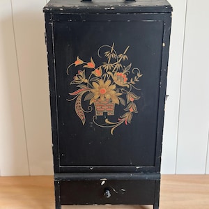 May include: A tall, rectangular black wooden cabinet featuring a top handle and a single drawer with a small knob. The front panel is decorated with a painted floral design in gold, orange, and red hues, showing signs of wear on the edges.