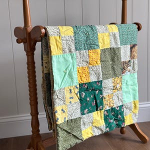 May include: A wooden quilt rack displaying a patchwork quilt. The quilt features squares of yellow, green, and patterned fabrics. The rack is made of light brown wood and has a decorative design.