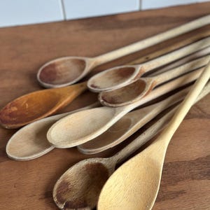 May include: A collection of multiple wooden spoons with varying shades of light and dark brown wood grain. The spoons feature long handles and oval-shaped bowls, showing signs of wear and natural texture across their surfaces.
