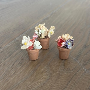 May include: Three miniature terracotta pots, each filled with colorful, handcrafted flowers. The flowers feature white, red, and purple petals with yellow centers. The pots are arranged on a wooden surface.
