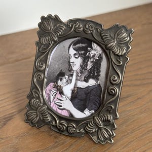 May include: Ornate, antique-style picture frame with a grayscale portrait of a woman holding a cat. The frame is a dark, metallic color with decorative floral and scrollwork details. The woman in the portrait is wearing a dark dress and a pink shawl.