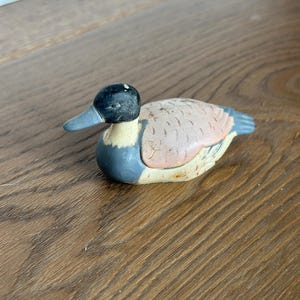 May include: A small, hand-painted wooden duck figurine. The duck has a black head, a light blue beak, and a body with shades of tan, blue, and yellow. The figurine is resting on a wooden surface.