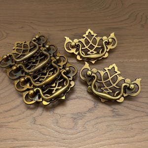 May include: A collection of antique brass drawer pulls. The pulls have a decorative, openwork design with a curved handle and ornate details. The hardware is arranged on a wooden surface.