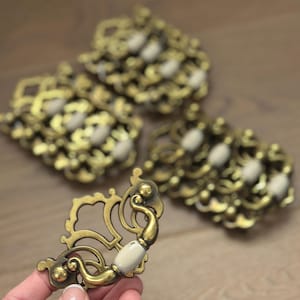 May include: Ornate brass drawer pulls with cream-colored ceramic handles. The pulls have a vintage aesthetic with intricate scrollwork and decorative details. The hardware is shown in a group of four.