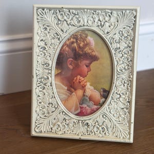 May include: Off-white, ornate picture frame with an oval portrait of a child. The frame has a detailed, embossed floral design. The portrait depicts a young child with blonde hair, hands clasped in prayer, and a small bird.