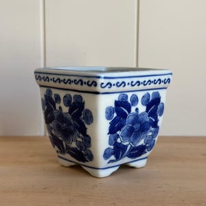 May include: A square ceramic planter featuring a white base with blue floral patterns and berries. The rim is decorated with a repeating blue scroll design. The planter stands on four small feet and has a tapered shape with a wide opening.
