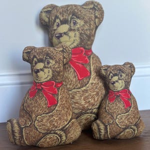 May include: Three stuffed teddy bear pillows of varying sizes, each featuring a brown textured fur print and a bright red ribbon tied around the neck. The bears are depicted in a seated position with visible facial features including eyes and noses.