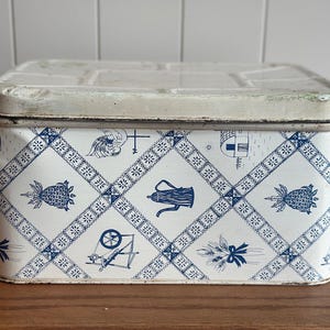 May include: A rectangular metal storage tin with a white base and a blue diamond-patterned grid. The design features illustrations of a coffee pot, a spinning wheel, a pineapple, and a weather vane within the grid sections. It has a lid.