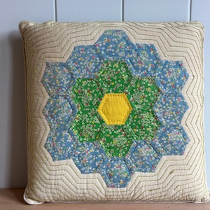 May include: A square throw pillow featuring a central hexagon patchwork design. The center is a solid yellow hexagon surrounded by green and blue floral-patterned hexagons. The background is cream fabric with wavy green stitched lines.