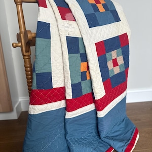 May include: A vintage quilt draped over a wooden chair. The quilt features a patchwork design with squares of blue, red, orange, and green, separated by white borders. The edges are trimmed with red binding. The quilt is a mix of colors and textures.