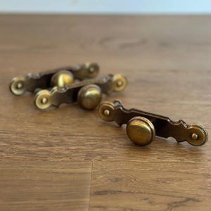 May include: Four antique brass drawer pulls with a decorative design. Each pull features a central knob and ornate detailing on the ends. The pulls are arranged on a wooden surface.