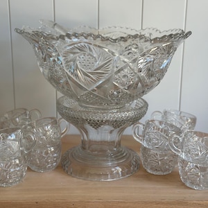 May include: A clear glass punch bowl set featuring a large, scalloped-edge bowl with intricate starburst and diamond-cut patterns, resting on a matching pedestal base, accompanied by several small, handled glass cups with similar cut-glass designs.