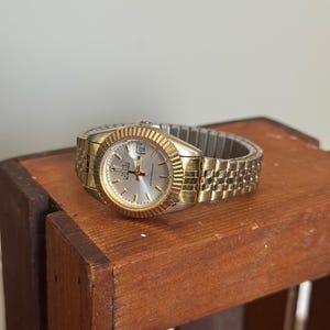 May include: A gold-toned wristwatch with a silver face and a date display at the 3 o'clock position. The watch has a textured bezel and a gold-toned metal band. The watch is water resistant.