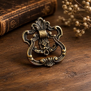 May include: Ornate antique brass drawer pull with a decorative, scrollwork design. The handle is a loop shape, and the pull has a detailed, vintage aesthetic. The brass finish has a warm, aged appearance, suggesting a classic or vintage style.