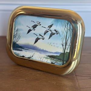 May include: A rectangular, gold-toned picture frame with a watercolor painting of geese in flight over a winter landscape. The painting features a blue sky, bare trees, and a snowy ground. The frame has rounded corners.
