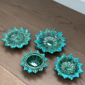 May include: Four turquoise and green ceramic bowls with a floral design. The bowls are arranged on a wooden surface. The bowls have a starburst shape with a detailed, textured pattern.