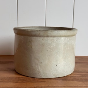 May include: A cylindrical ceramic crock with a light beige, off-white finish and a smooth, slightly textured surface. The vessel features a thick, rounded rim at the top and a flat base, presenting a simple, utilitarian design with a neutral tone.