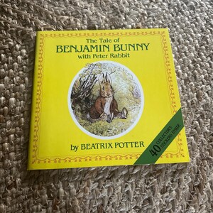 May include: A children's book titled "The Tale of Benjamin Bunny with Peter Rabbit" by Beatrix Potter. The cover is yellow with a circular illustration of a rabbit. Includes 40 collector's stickers.