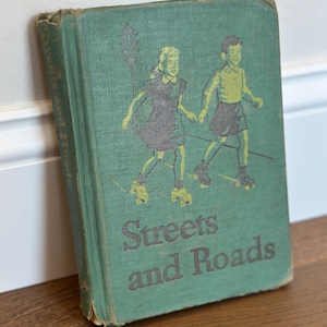 May include: A vintage green hardcover book titled Streets and Roads. The front cover features a dark gray and yellow illustration of two children wearing roller skates, standing near a street lamp. The book shows signs of wear on the edges.
