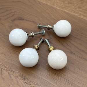 May include: Four white ceramic ball cabinet knobs with metal hardware. The knobs are round and smooth, with a glossy finish. The hardware includes a threaded screw for installation.