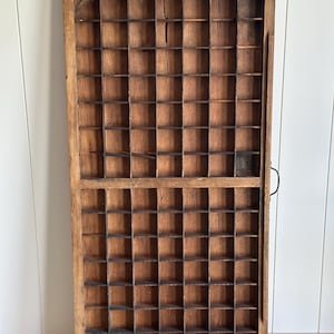 May include: A rectangular wooden printer's tray featuring a grid of numerous small, open compartments. The natural brown wood shows visible grain and texture, with a metal handle attached to the right side of the frame for easy handling.