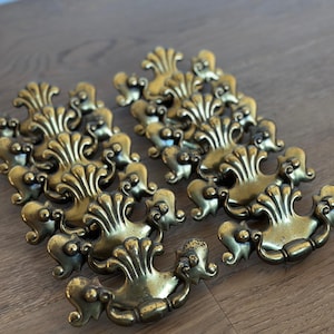 May include: A collection of antique brass drawer pulls with ornate, symmetrical designs. Each pull features a central fan-shaped element flanked by scrolling details and small, rounded accents. The hardware has a warm, aged patina, suggesting vintage charm.