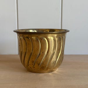 May include: A polished brass planter featuring a rounded shape and a series of vertical, swirling fluted grooves along the exterior surface. The metal has a warm, golden tone with a smooth rim and a slightly tapered base, showing natural patina.