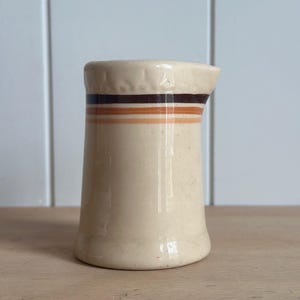 May include: A cream-colored ceramic creamer pitcher featuring a cylindrical body with a slightly flared base and a small pouring spout. The upper section is decorated with three horizontal stripes in dark brown, medium brown, and light orange.