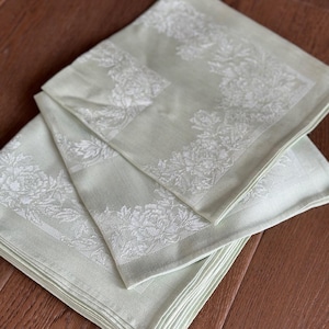 May include: A stack of pale green cloth napkins with a white floral pattern. The napkins are folded and stacked on a wooden surface. The floral design is visible on the edges of the napkins.